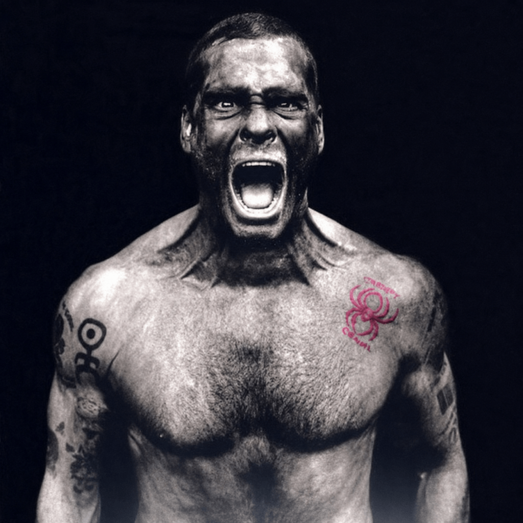 A muscular man with an intense expression, screaming. He has various tattoos on his arms and a spider tattoo on his shoulder, with a dark background.