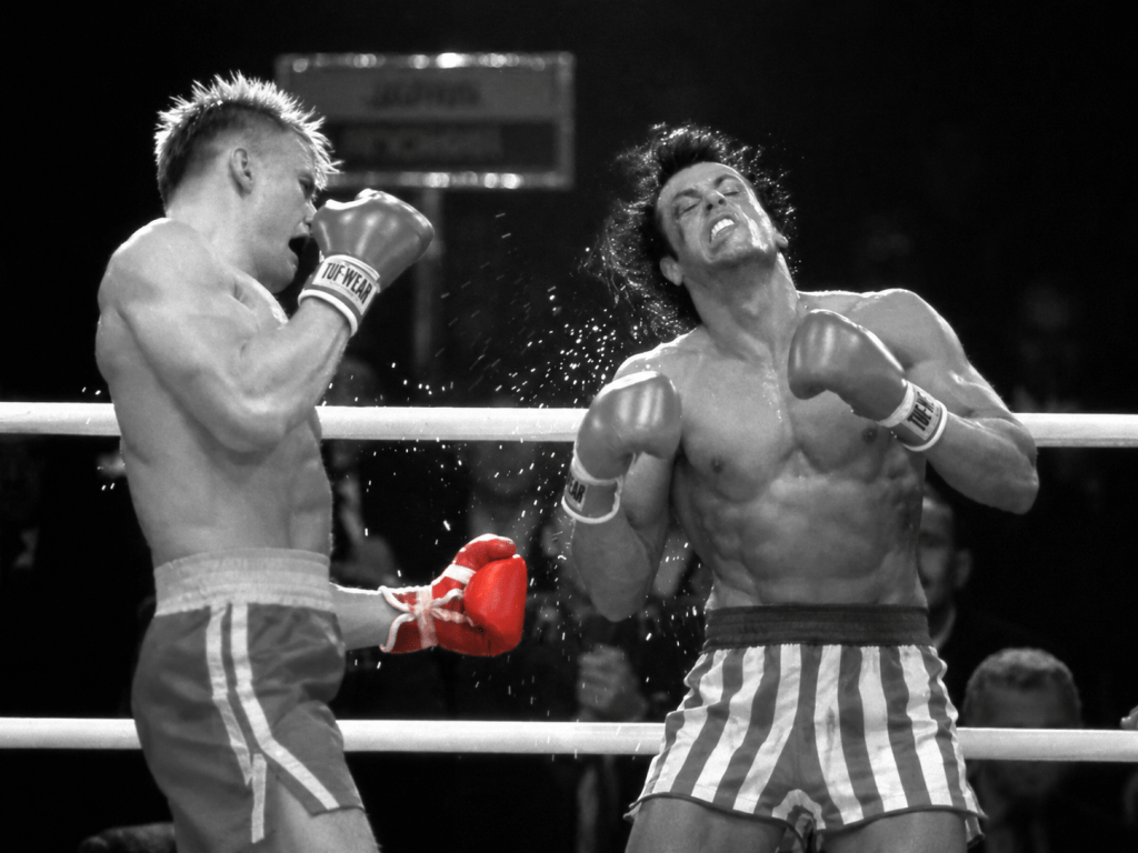 A dramatic boxing match scene featuring two male fighters in the ring. One fighter, wearing red gloves, appears to be delivering a powerful punch, while the other is bracing for impact, showing a pained expression.