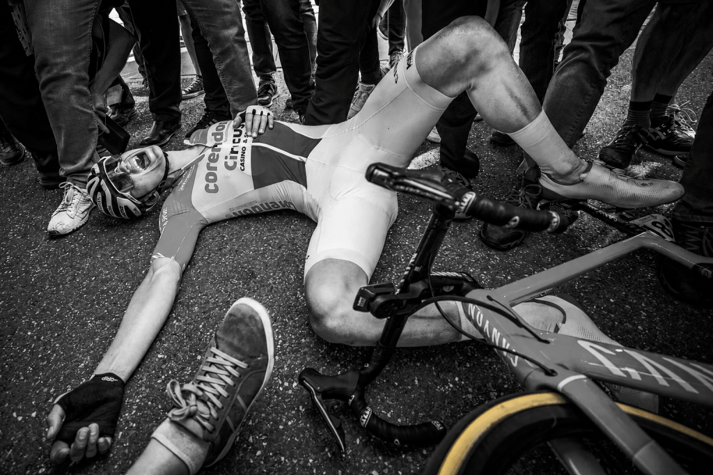 A cyclist lying on the ground, appearing exhausted and in pain, surrounded by spectators. The scene captures the intensity of a cycling event in black and white.