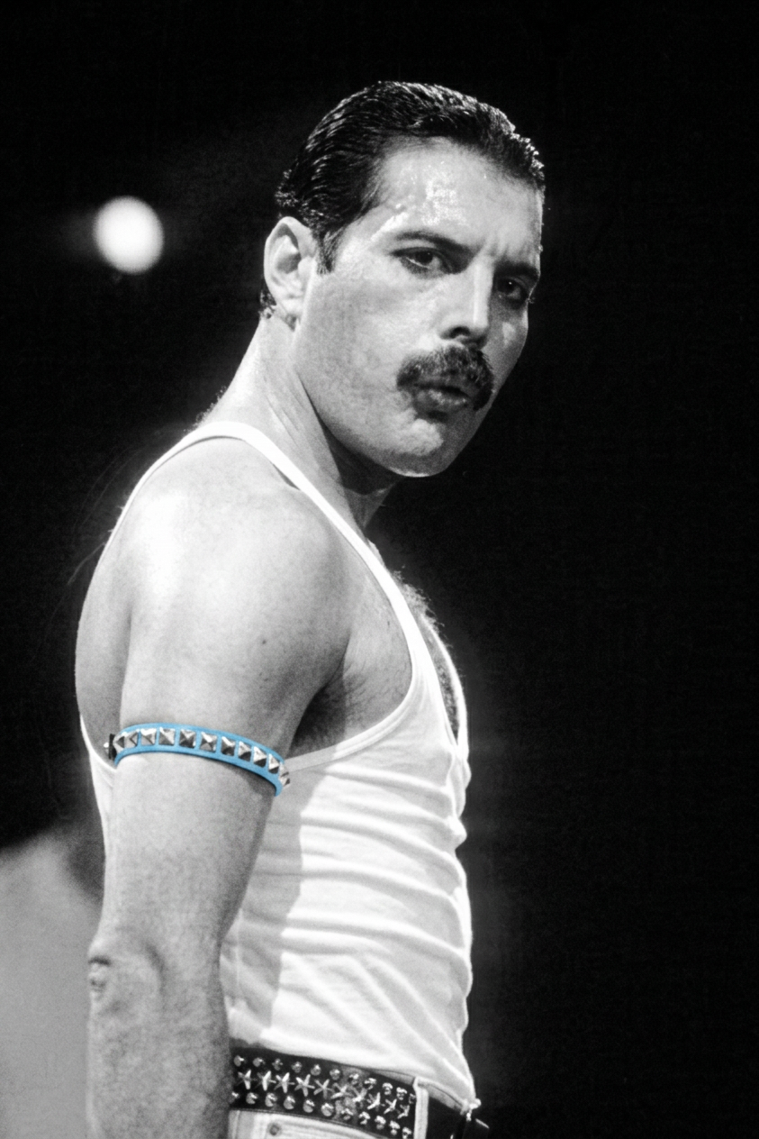 A black and white image of a male singer wearing a white tank top and a studded belt, posing confidently with a serious expression. He has a dark mustache and an armband decorated with blue studs on his upper arm.