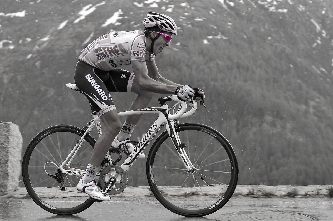 A cyclist in a racing outfit riding a white road bike on a wet road with mountains in the background.