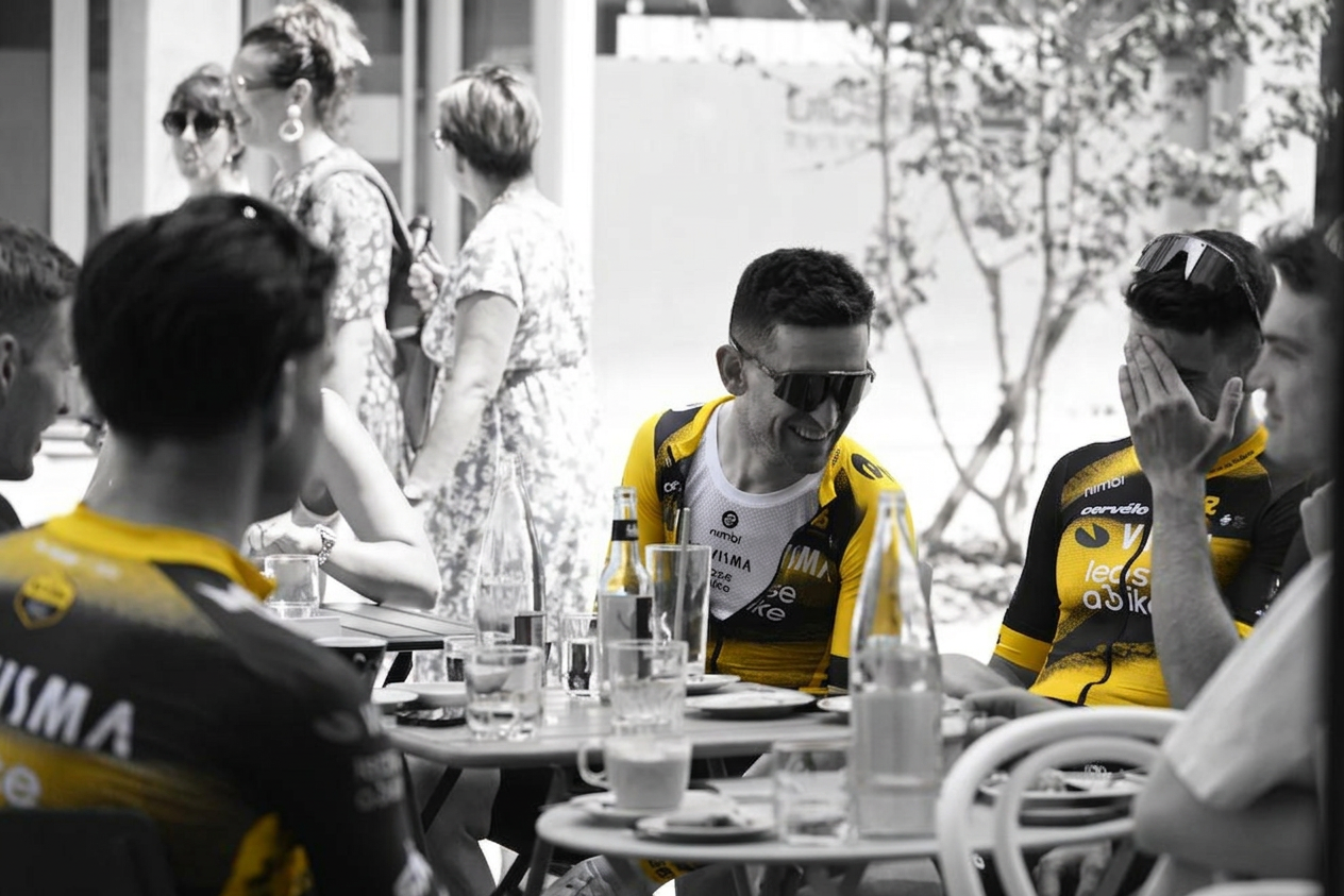 A group of cyclists in yellow jerseys laughing and enjoying drinks at an outdoor cafe, with blurred patrons in the background.