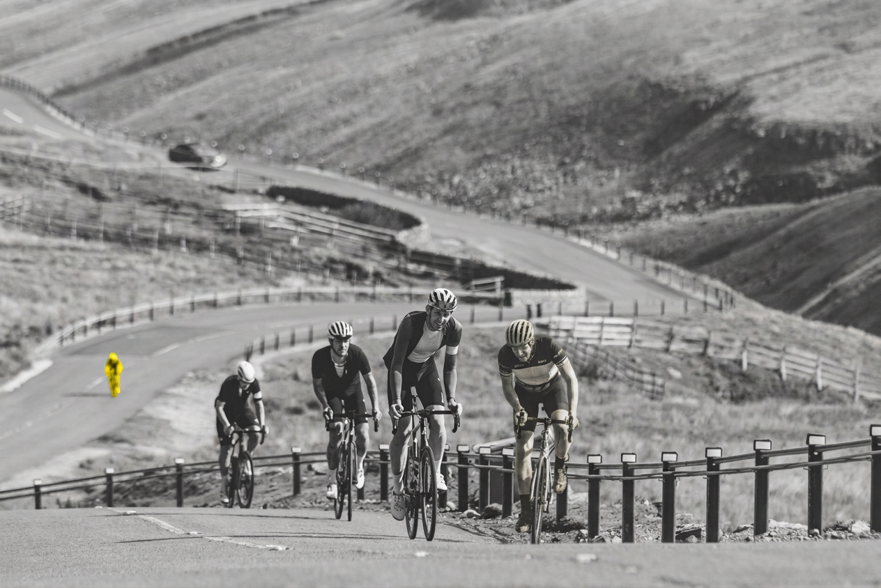 A group of four cyclists riding uphill on a winding road, with a bright yellow figure in the background. The landscape is mostly in shades of gray, emphasizing the cyclists' effort and the road's curves.