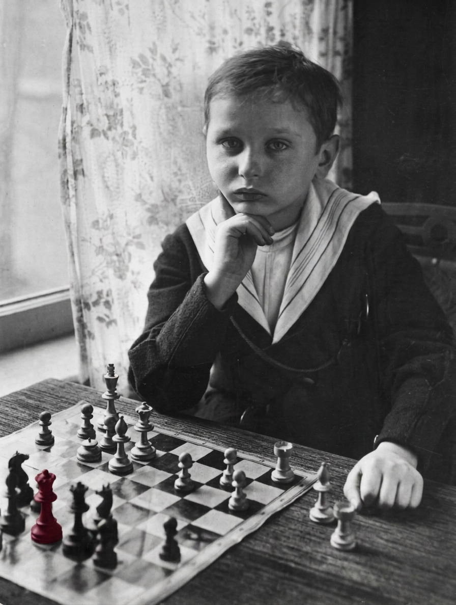 A young boy in a sailor-style outfit sits thoughtfully at a chessboard, with a red chess piece prominently displayed among other pieces, suggesting deep concentration on the game.
